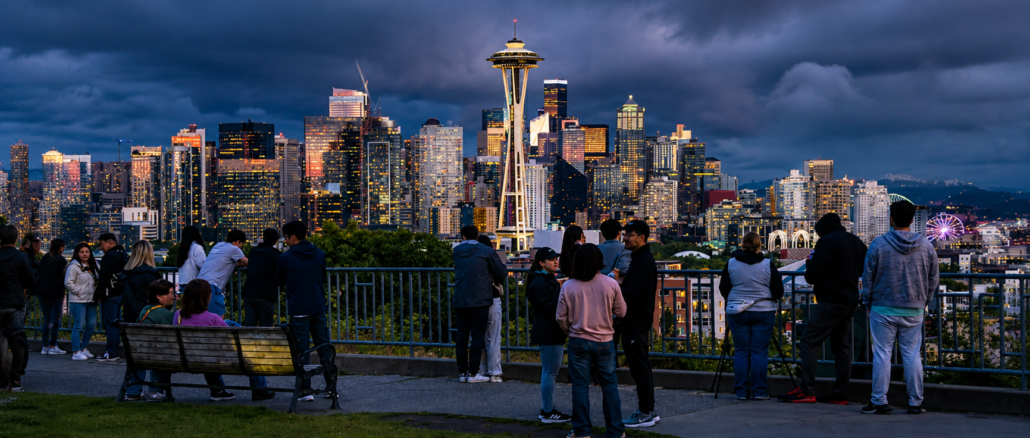 Vista panorámica del centro de Seattle al atardecer con la Space Needle y personas observando el horizonte desde un mirador urbano.