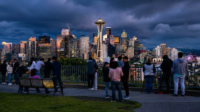 Vista panorámica del centro de Seattle al atardecer con la Space Needle y personas observando el horizonte desde un mirador urbano.